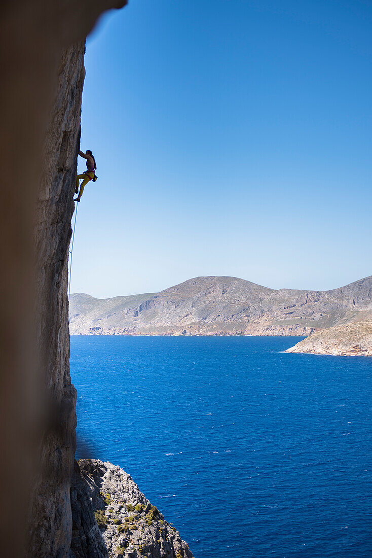 Woman climbing steep cliff in scenery with sea and coastline, Kalymnos, Greece