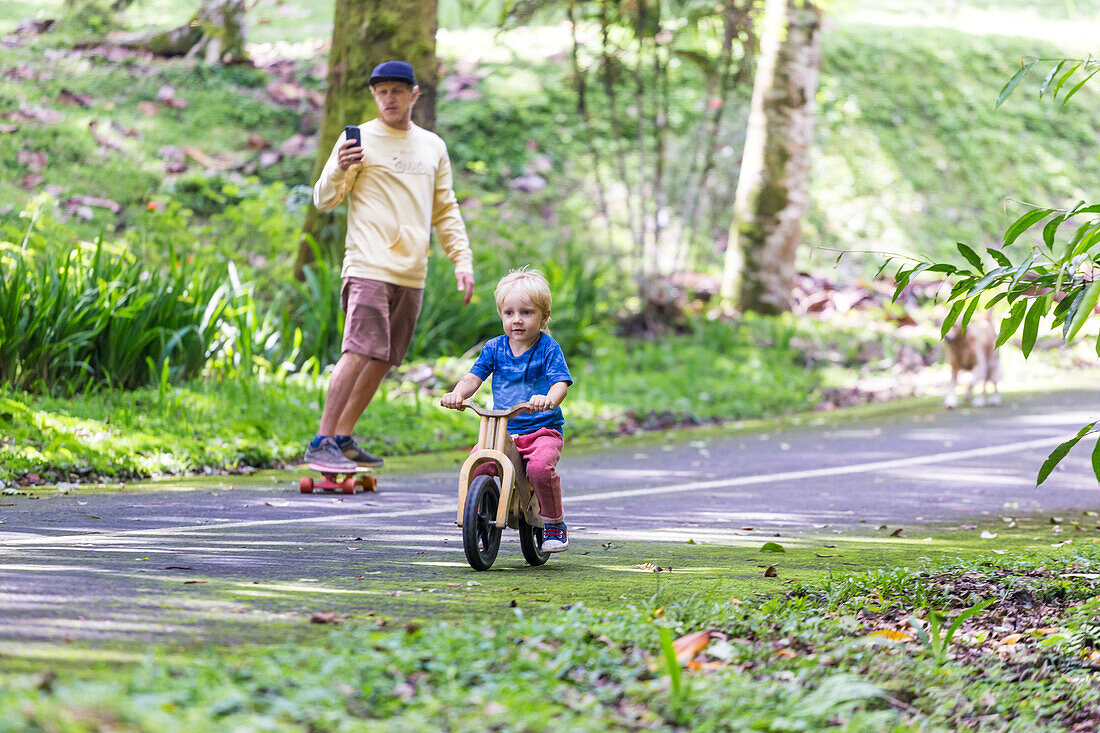 Father on skateboard with phone accompanying son on bicycle
