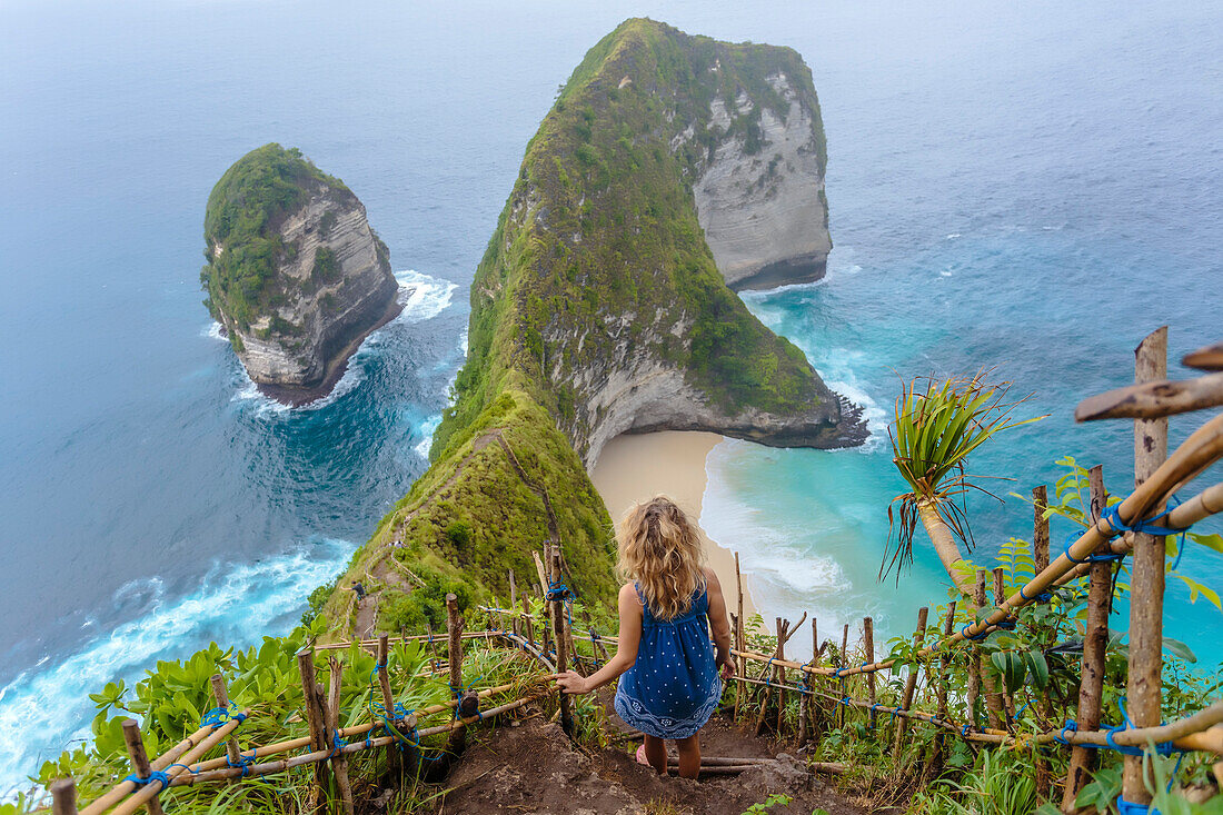 Blonde woman standing on cliffside steps admiring ocean coastline of Nusa Penida island, Bali, Indonesia