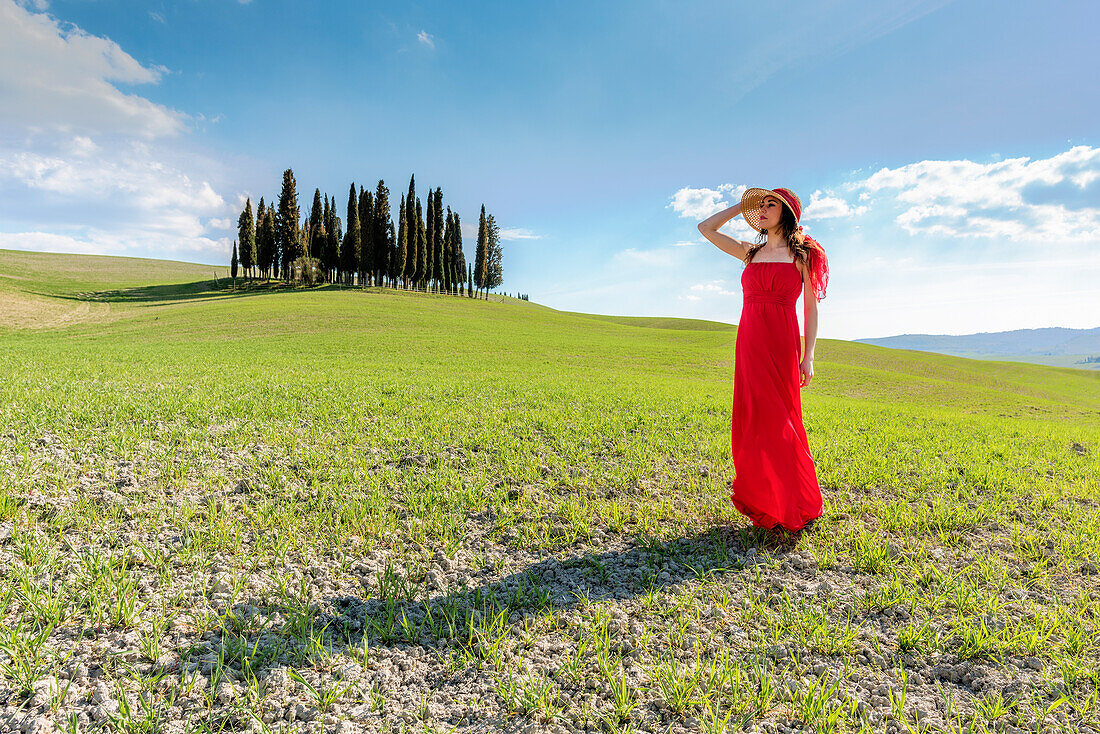 Eine junge Frau in rotem Kleid bewundert die Aussicht in einem Weizenfeld in der Nähe der Zypressen im Orcia-Tal