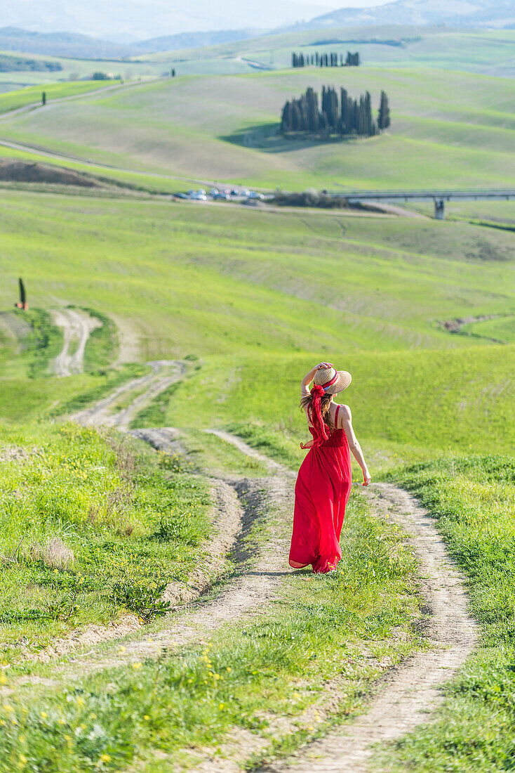 San Quirico d'Orcia, Orcia valley, Siena, Tuscany, Italy. A young woman in red dress walking along a country path near the cypresses of val d'orcia