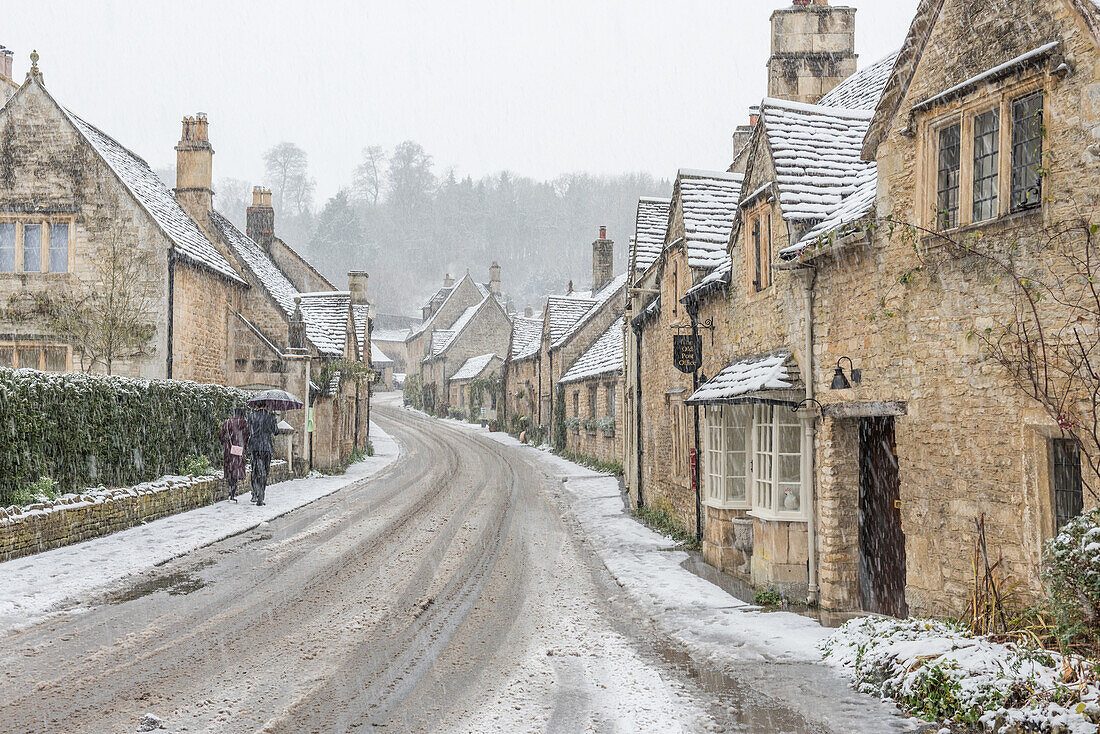 A couple walking up the hill in Castle Combe with an umbrella protecting them from the heavy snowfall, Wiltshire, England, United Kingdom, Europe