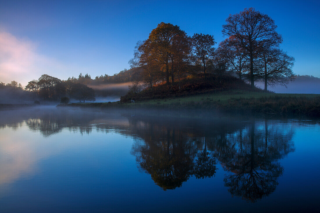 A calm clear autumn morning at Elterwater as the first light of a new day illuminates the trees and mist, Lake District National Park, UNESCO World Heritage Site, Cumbria, England, United Kingdom, Europe