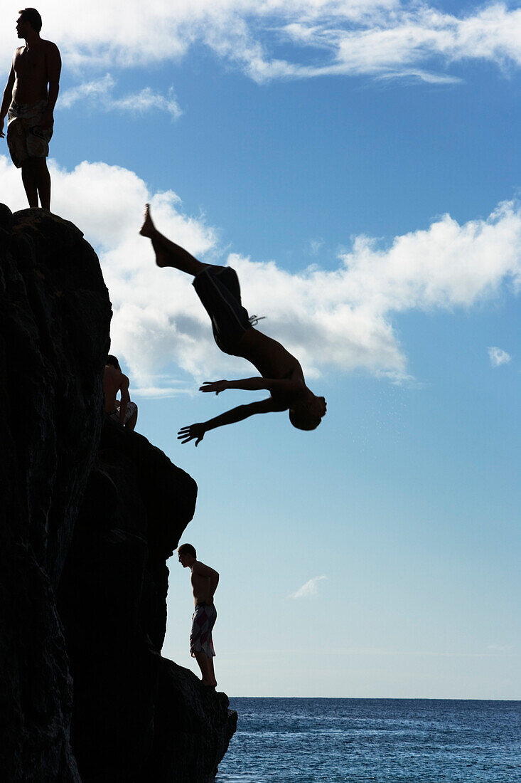 Silhouetted young men and a man upside down mid-air, jumping off of the 'da Big Rock' or 'Jump Rock', a rock that is perfectly positioned in the bay at Waimea Bay Beach Park, O'ahu, Hawaii.