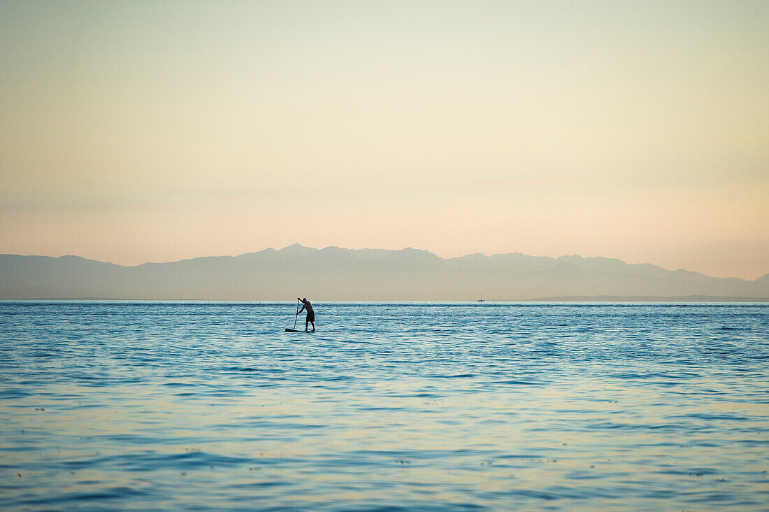 Standing up paddle boarder paddling on Vancouver's English Bay at dusk, British Columbia, Canada