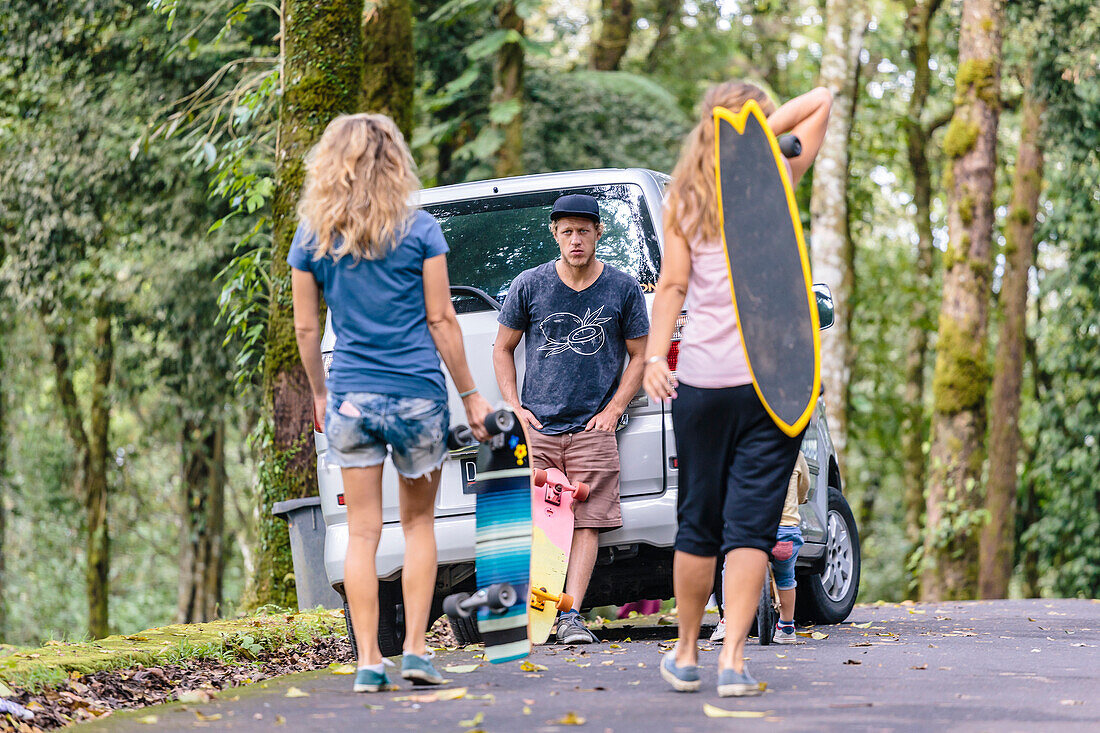 Two women carrying skateboards walking towards male friend waiting and leaning on parked car, Bali, Indonesia