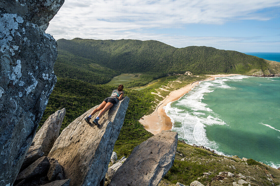 Young woman lying on edge of rock with view from Pico da Coroa Hill to Lagoinha do Leste Beach, Florianopolis, Santa Catarina Island, Brazil