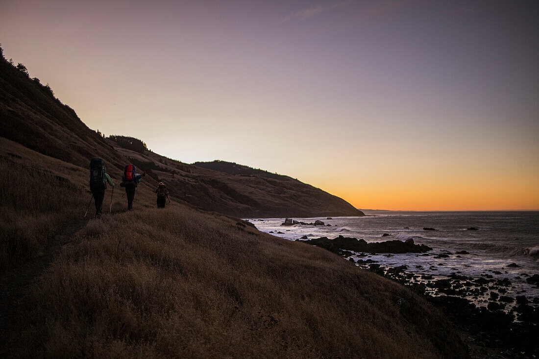 Three backpackers hiking along shore of Lost Coast at dawn, California, USA