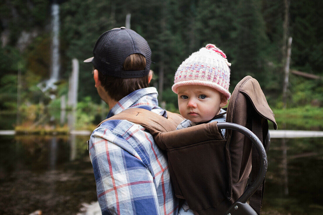 Baby girl sitting on fathers back in backpack carrier and looking at camera during forest hike, Washington, USA
