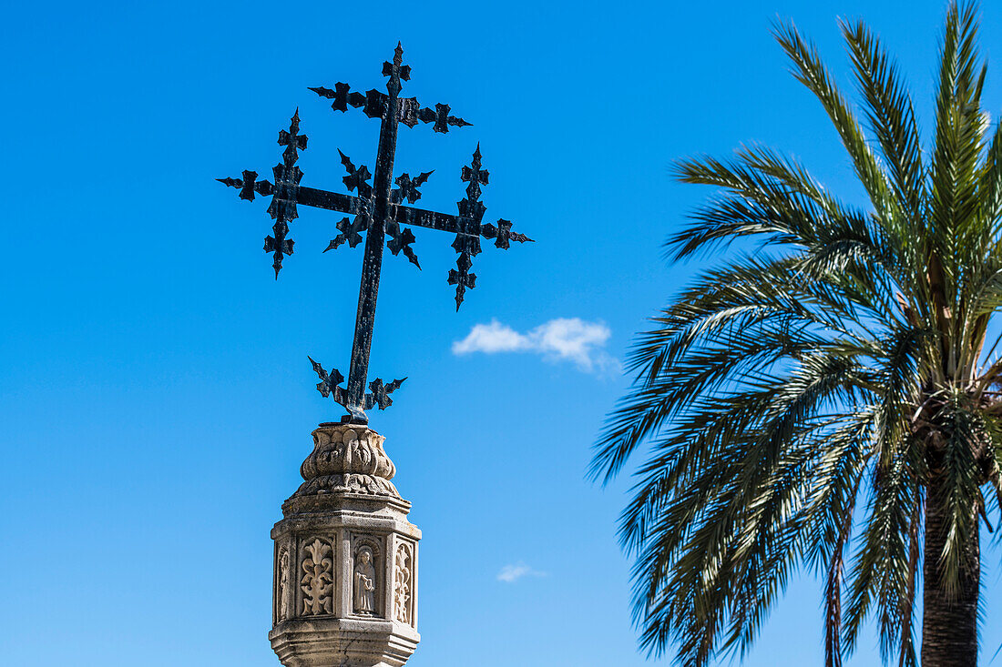 Ein Kreuz auf einer Steinsäule neben einer Palme vor der Kirche, Selva, Mallorca, Spanien