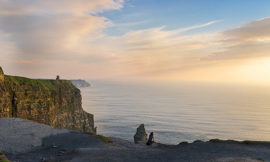 Cliffs of Moher, Liscannor, Co. Clare, Munster province, Ireland. A woman watch the sunset sitting on a rock.
