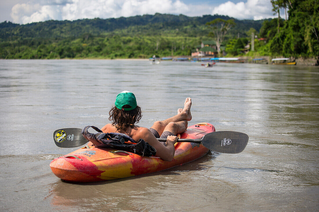 Young man relaxes in whitewater kayak on the Alto (Upper) Madre de Dios river int he jungles of Peru in the Peruvian Amazon. Village of Atalaya in the distance.
