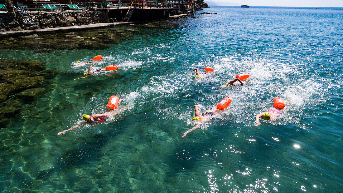 group of seven young swimmers - each with a life buoy - training in the clear and transparent waters of the Ligurian sea with terrace and pontoon behind them and a boat in the background on a sunny day in Paraggi, Cinque Terre, Italy