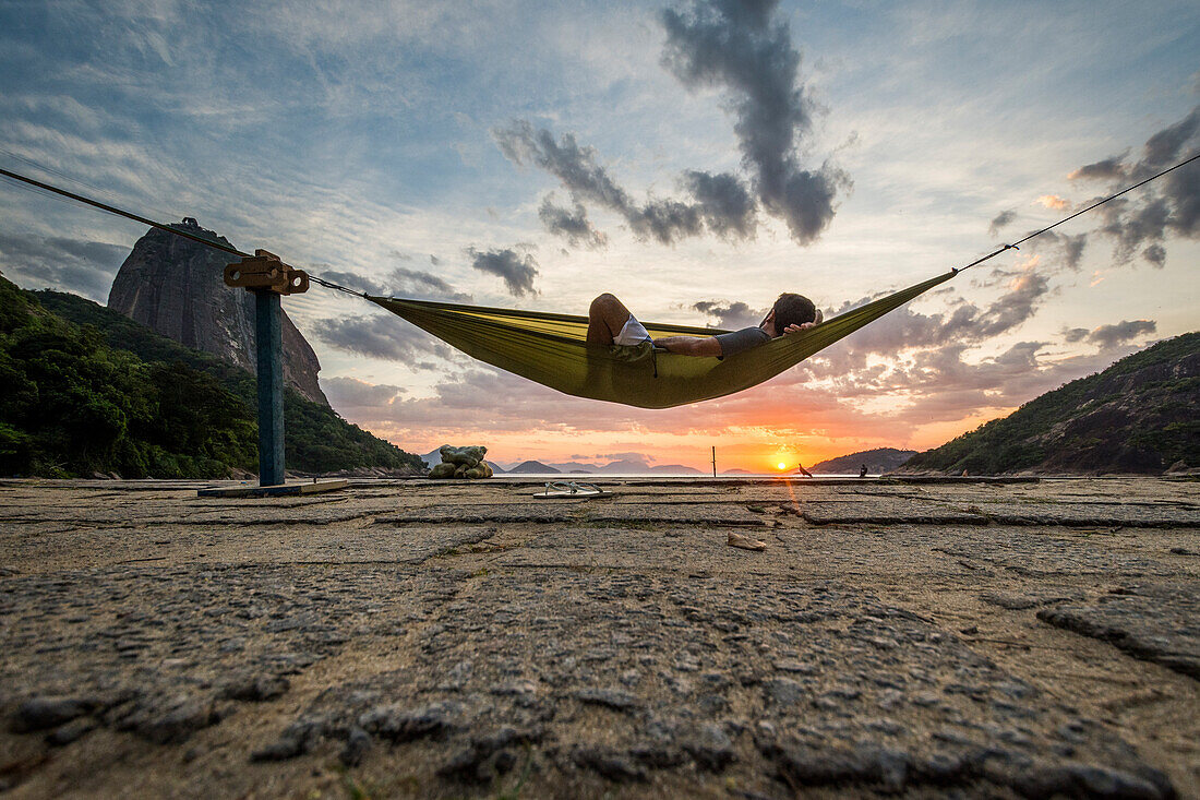 Man lying in hammock on Vermelha Beach at sunrise in Rio de Janeiro with Sugarloaf Mountain in background, Brazil