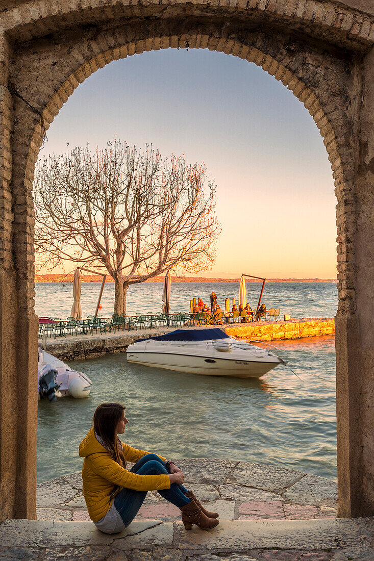 Tourist admiring the sunset light on the little harbour of Punta San Vigilio from the arc of the charming 16th-century inn on the eastern shore of Lake Garda, Verona province, Veneto, Italy.