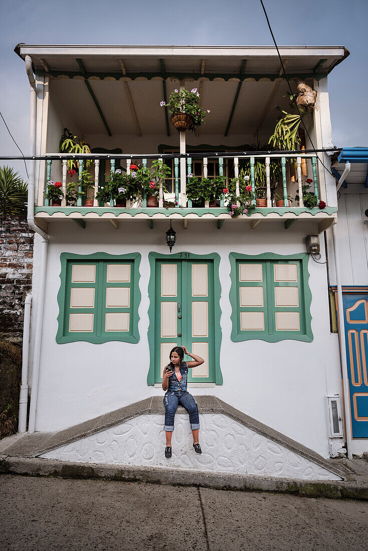 junge Kolumbianerin macht sich die Haare mit ihrem Smartphone vor einem kolonialen Gebäude, Salento, UNESCO Welterbe Kaffee Dreieck (Zona Cafatera), Departmento Quindio, Kolumbien, Südamerika