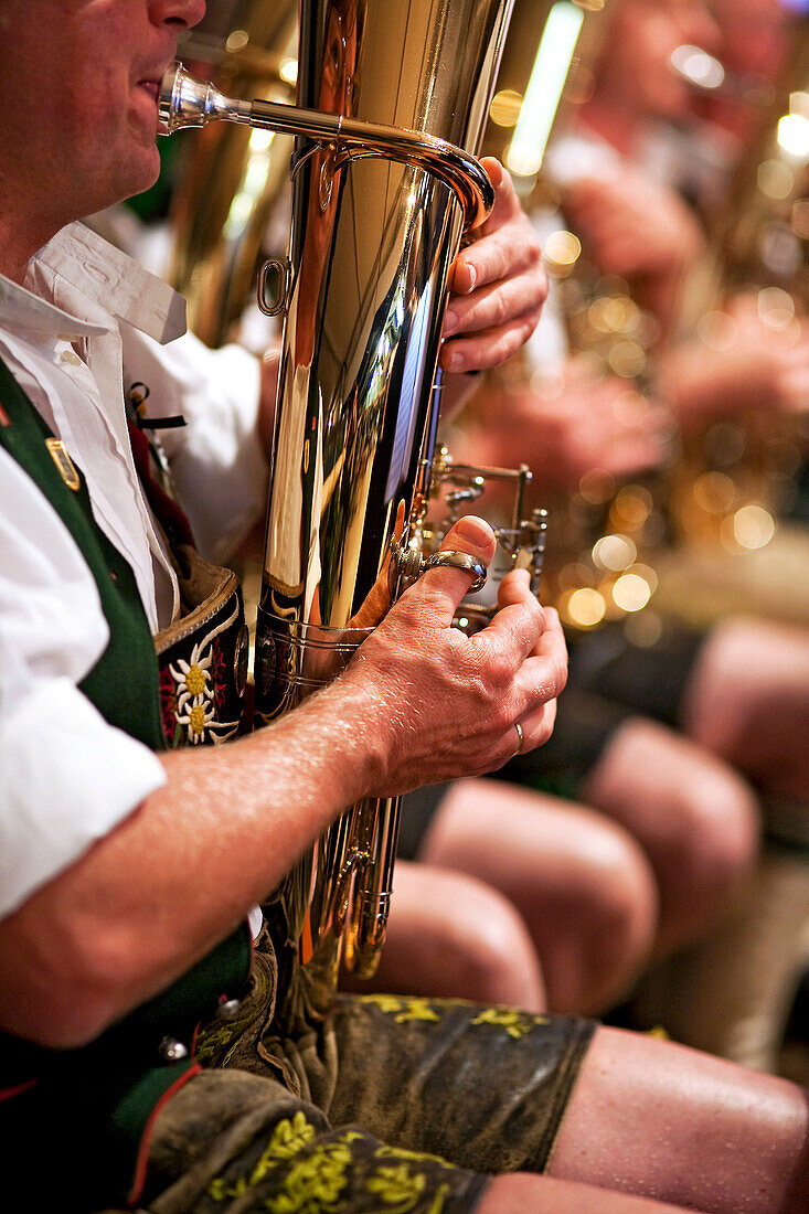 Musician in bavarian costume plays tenor horn; further musicians blurry in the background; in detail