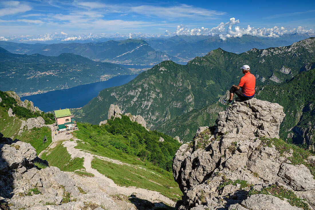 Mann beim Wandern sitzt auf Felsabsatz und blickt auf Rifugio Rosalba und Comer See, an der Grignetta, Grigne, Bergamasker Alpen, Lombardei, Italien