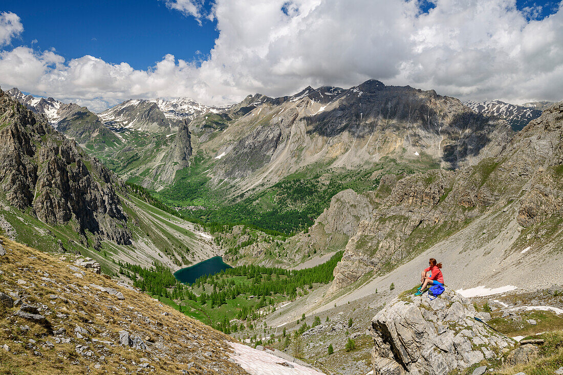 Frau beim Wandern sitzt auf Felsen und blickt ins Tal, Bergsee und Cottische Alpen im Hintergrund, Val Maira, Cottische Alpen, Piemont, Italien