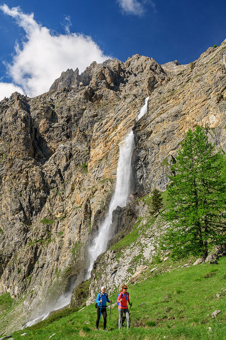 Man and woman hiking ascending towards hut rifugio Stroppia, waterfall cascata Stroppia in background, Val Maira, Cottian Alps, Piedmont, Italy