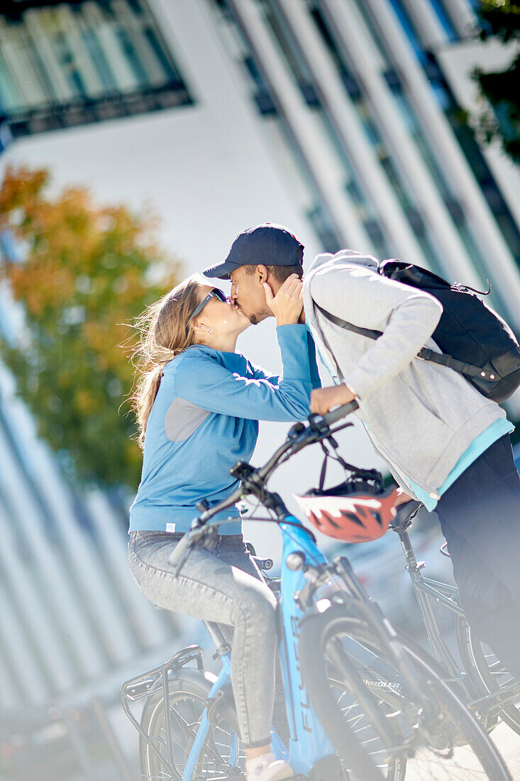 Young  woman kissing young man on eBikes downtown, Munich, bavaria, germany
