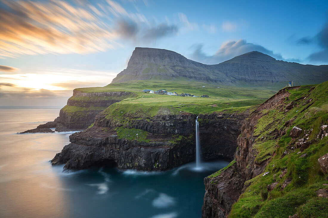 hiker contemplating the mountains, the cliffs and the waterfall cascading into the sea by the village of gasadalur, vagar, faroe islands, denmark