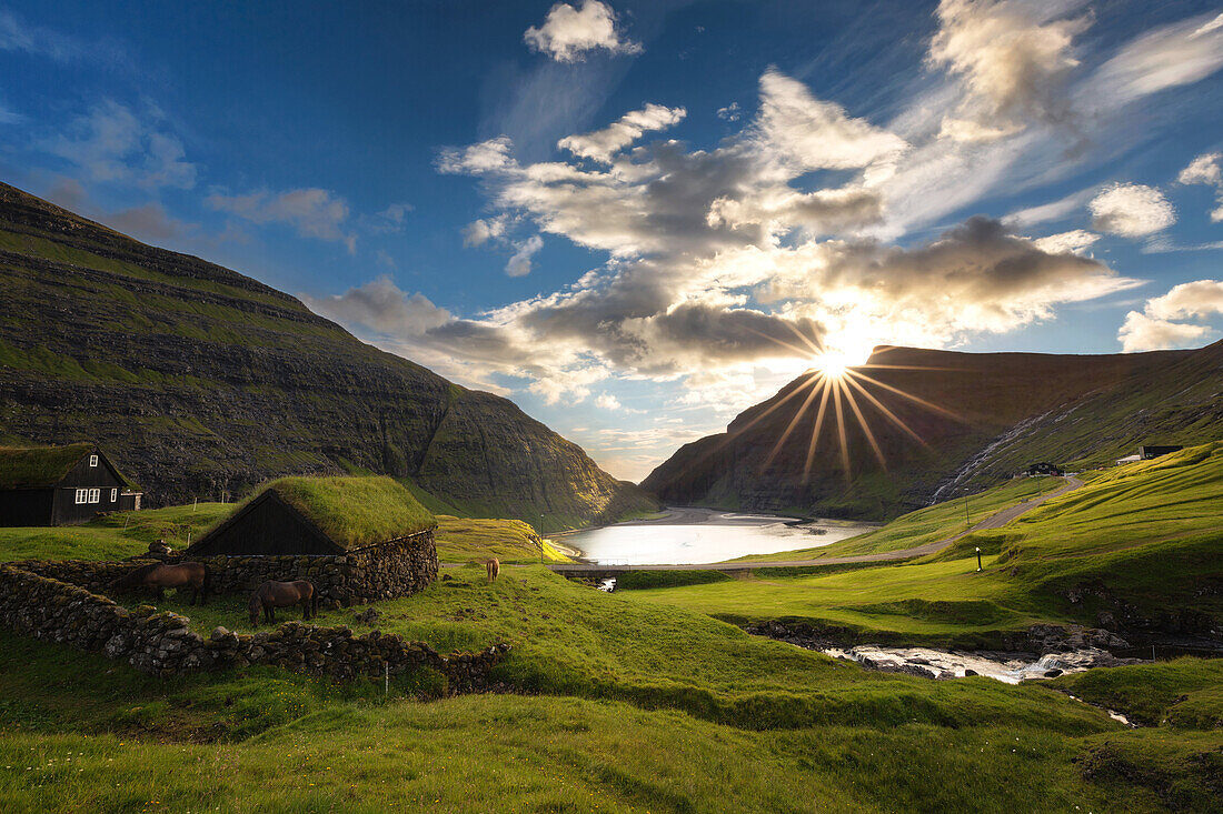 traditional houses with vegetal roofs in a verdant fjord, the last rays of the setting sun over the distant cliffs, saksun, streymoy, faroe islands, denmark