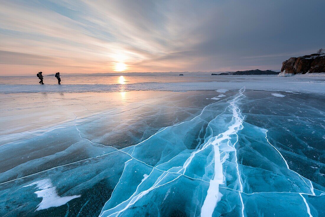 Two persons walking over a flat ice with cracks on the lake Baikal at sunrise, Irkutsk region, Siberia, Russia.