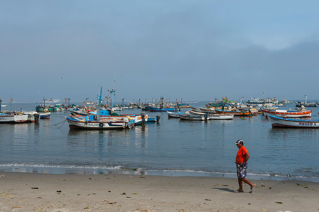 Ein Mann geht am Strand entlang vor einer Kulisse von Fischerbooten, Paracas, Ica, Peru, Südamerika