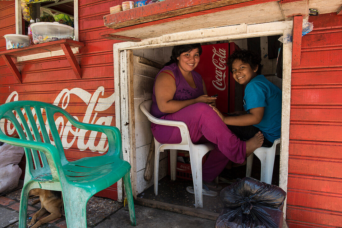 Eine Frau und ihr Sohn machen eine Pause vom Verkauf von Snacks und Getränken in einem kleinen Holzbau neben einer der Hauptstraßen der Stadt, Leticia, Amazonas, Kolumbien, Südamerika