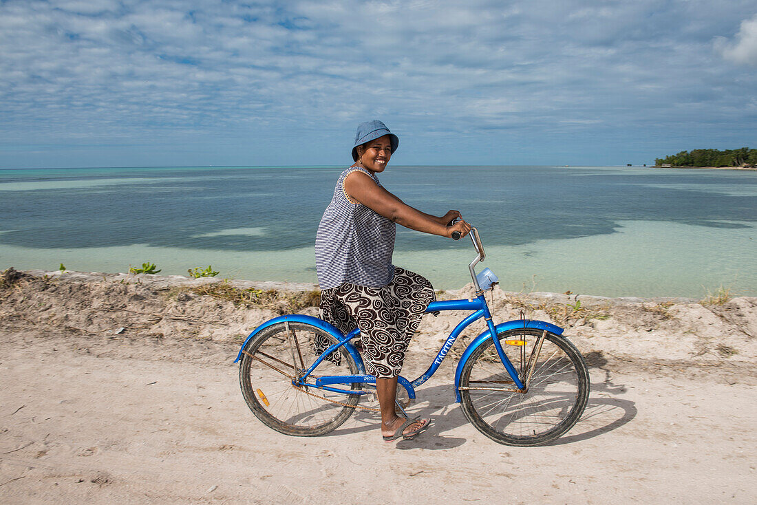 Eine Frau mit blauem Hut und langem Rock fährt ein blaues Fahrrad entlang des Wassers, Butaritari Atoll, Gilbert-Inseln, Kiribati, Südpazifik