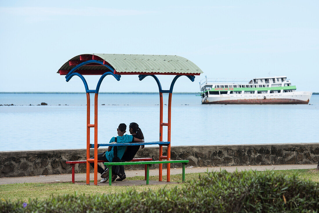 A young couple sits on a colorful resting place overlooking the shore, which features a boat leaning to port in shallow, low-tidal waters, Lautoka, Viti Levu, Fiji, South Pacific