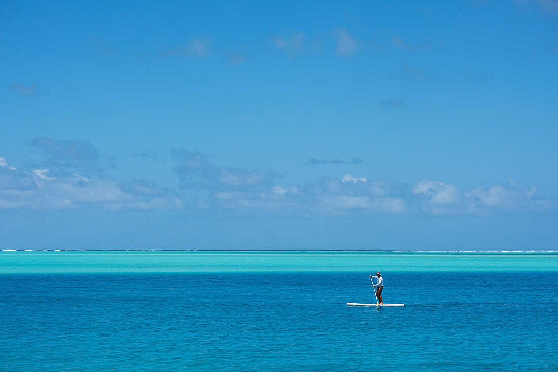 A lone standup paddle surfer wearing a long-sleeved shirt, a swimsuit, and a hat, paddles through dark-blue waters with shallower, lighter-colored water in the background, Huahine, Society Islands, French Polynesia, South Pacific