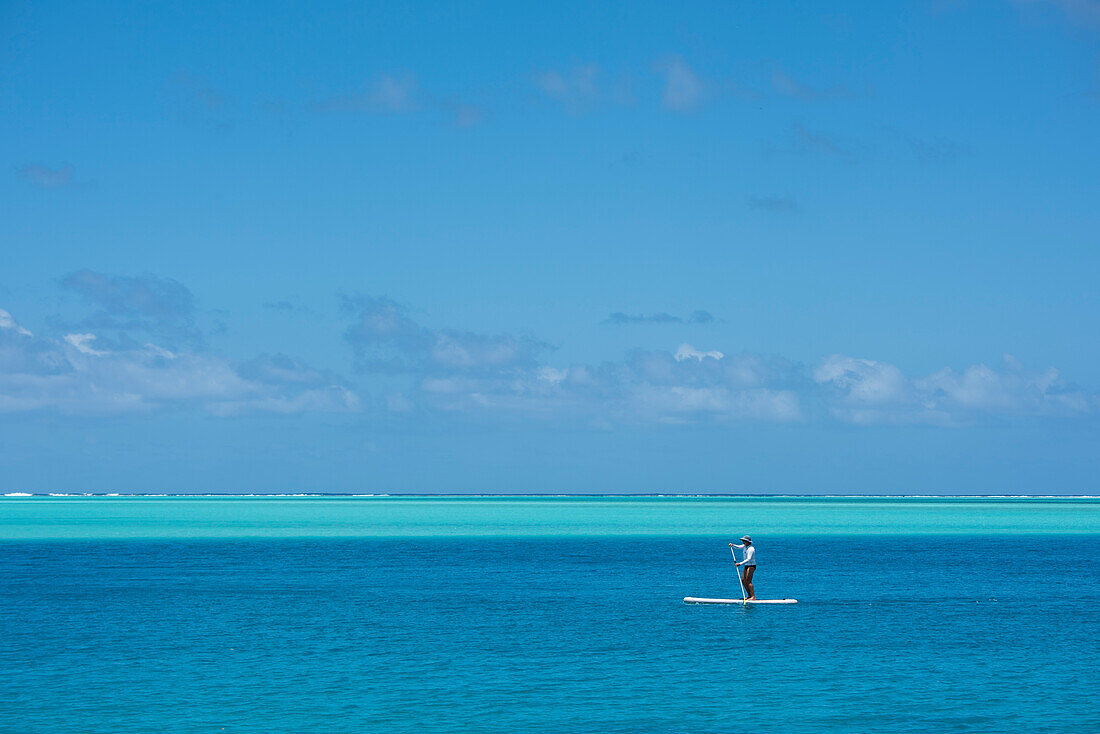 Ein einzelner Standup Paddler, der ein langärmeliges Hemd, einen Badeanzug und einen Hut trägt, paddelt durch dunkelblaues Wasser, im Hintergrund ist das Wasser flacher und heller, Huahine, Gesellschaftsinseln, Französisch-Polynesien, Südpazifik