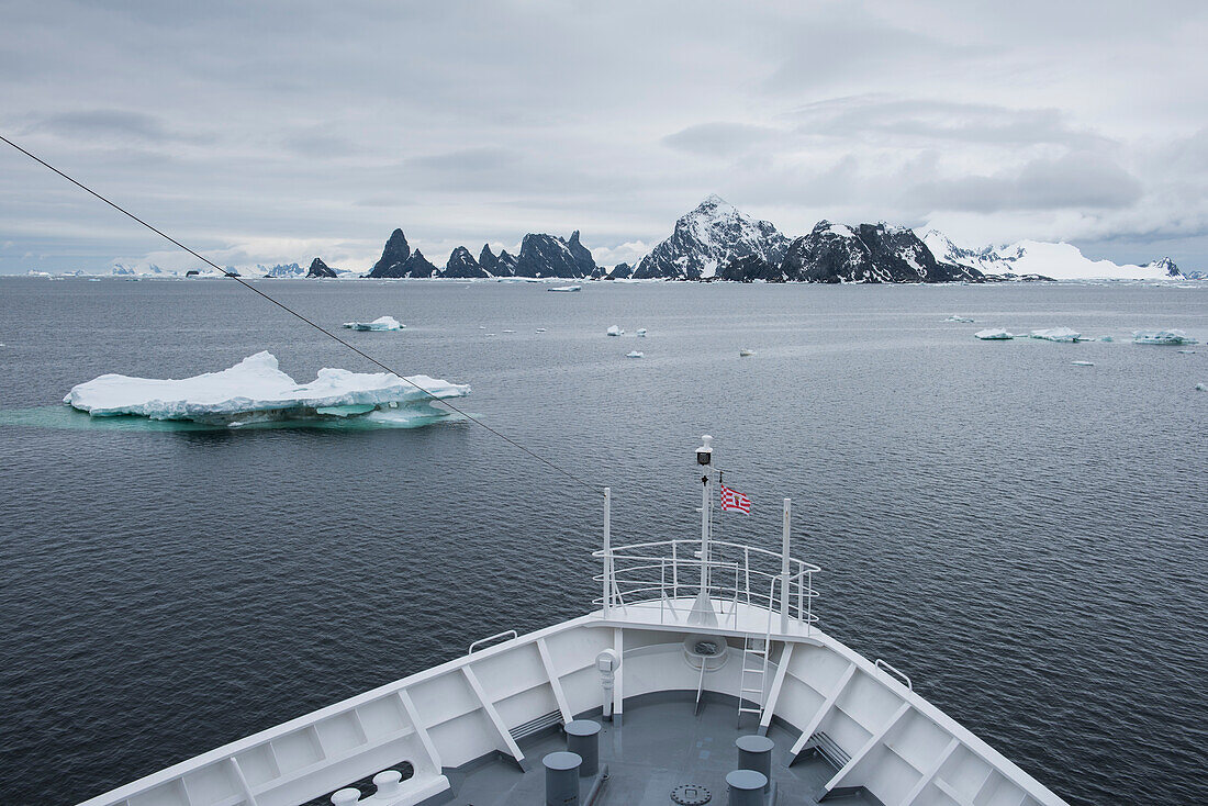 The bow of expedition cruise ship MS Bremen (Hapag-Lloyd Cruises) is visible as the vessel approaches a craggy, mountainous island surrounded by several ice-floes, Laurie Island, South Orkney Islands, Antarctica