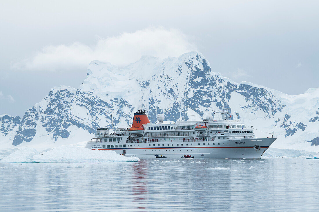 Expedition cruise ship MS Bremen (Hapag-Lloyd Cruises), seen beneath a large, snow-covered mountain, sends Zodiac dinghy rafts with passengers on a scenic cruise around the bay, Paradise Bay (Paradise Harbor), Danco Coast, Graham Land, Antarctica