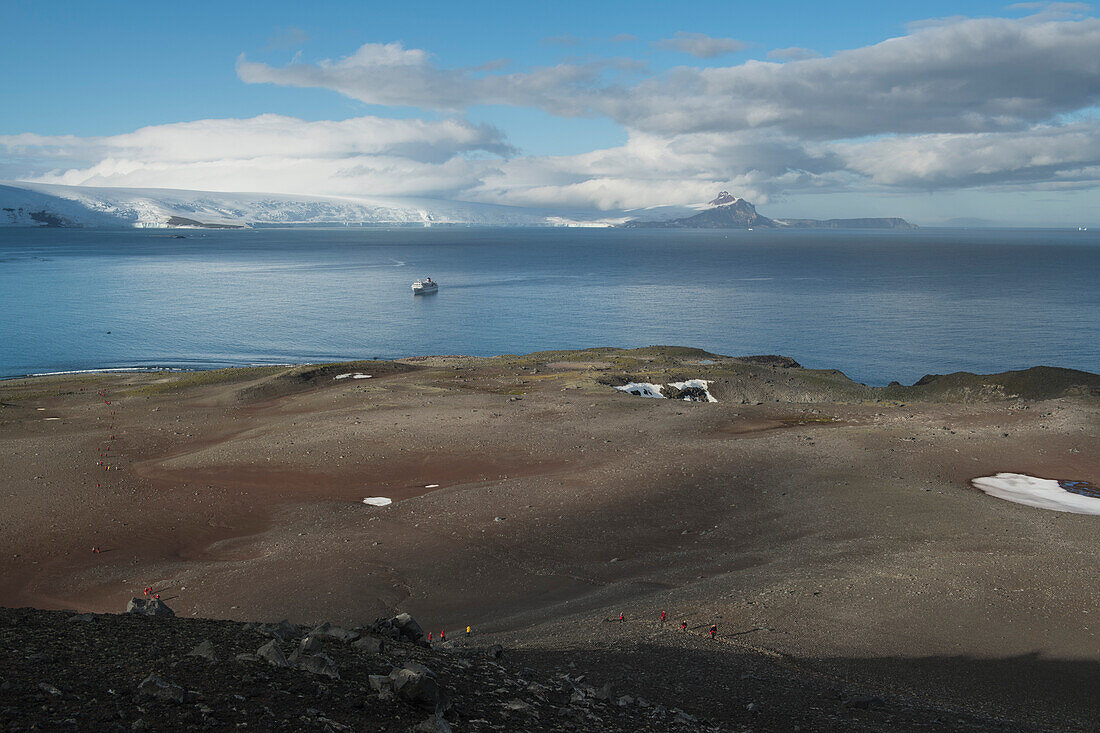 Das Expeditions-Kreuzfahrtschiff MS Bremen (Hapag-Lloyd Kreuzfahrten) liegt vor Anker, während die Passagiere im Vordergrund eine breite felsige Ebene durchqueren, Penguin Island, Südshetland-Inseln, Antarktis