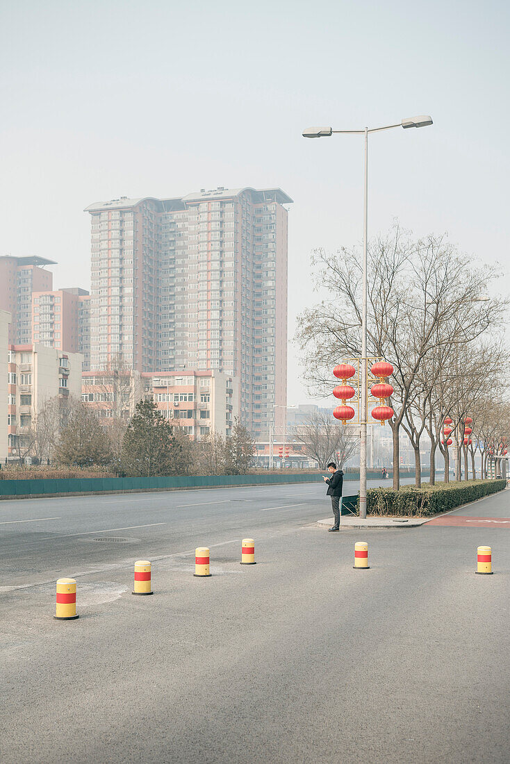 Chinese steht an leerer Straße und blickt auf Handy, Kongresszentrum (CNCC) im Smog der Großstadt, Peking, China, Asien