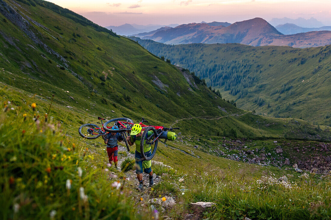 Zwei Mountainbiker tragen ihre Räder nach Sonnenuntergang auf einen Berg, grüne Bergwiese, Kirchberg, Tirol, Österreich