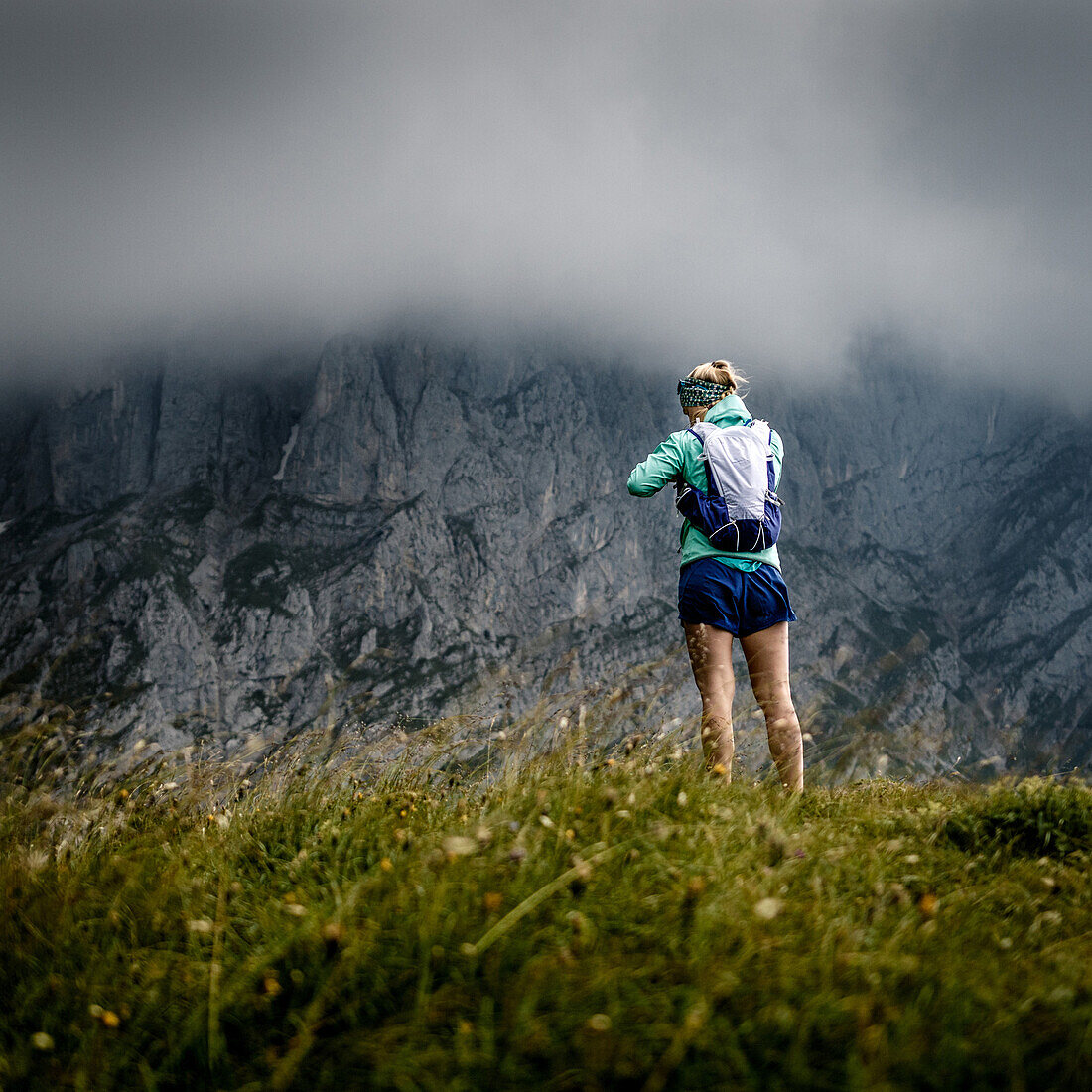 Junge Frau mit Laufbekleidung steht auf einem grasigen Buckel vor den Felswänden des Wilden Kaisers, Stürmisches Wetter,Scheffau, Tirol, Österreich