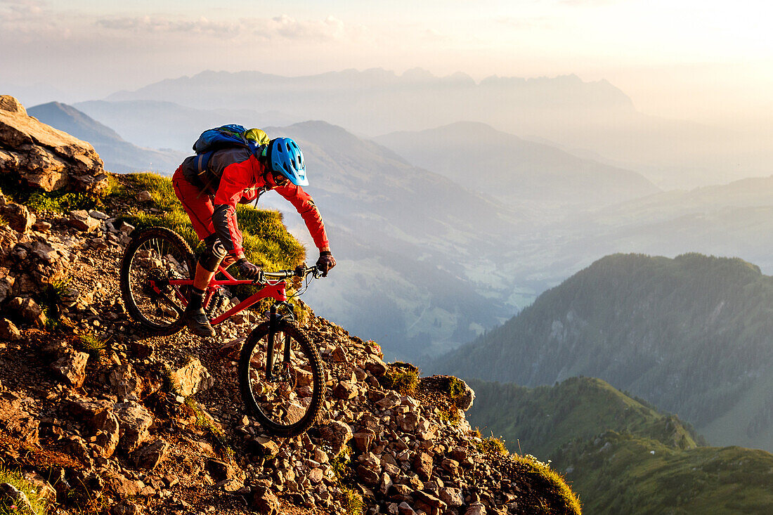 Middle aged man rides down a rocky and steep hiking path on his mountainbike, morning light, Wilder Kaiser mountain range in the background, Kirchberg, Tyrol, Austria