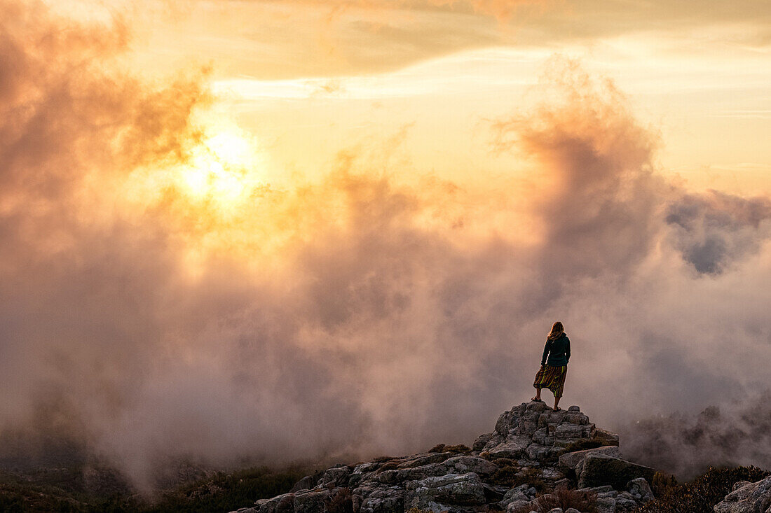 Young woman on a rock, sunrise with dramatic cloud scenery, Corsica