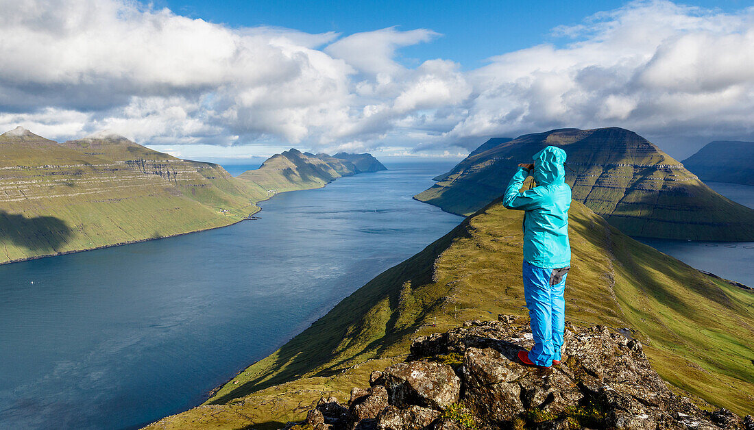 Young woman looks over the fjord landscape, the mountains and the ozean with a pair of binoculars on a bluebird day, Faroe Islands