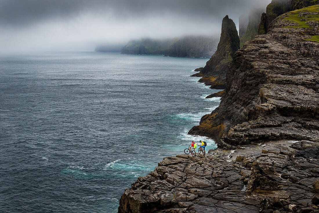 Two mountainbikers standing on a rock clif at the Atlantic Ocean, Faroe Islands