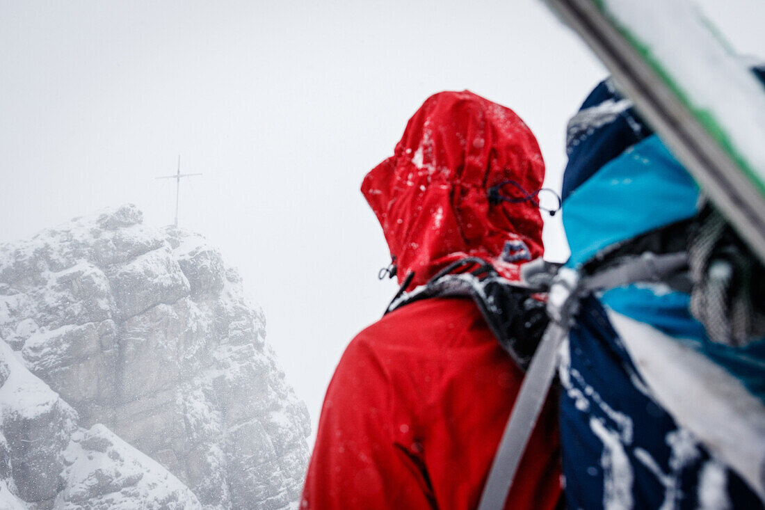 Young ski mountaineer has a look at the summit cross of mount Ponten, snowfall and mist, Allgäu, Germany