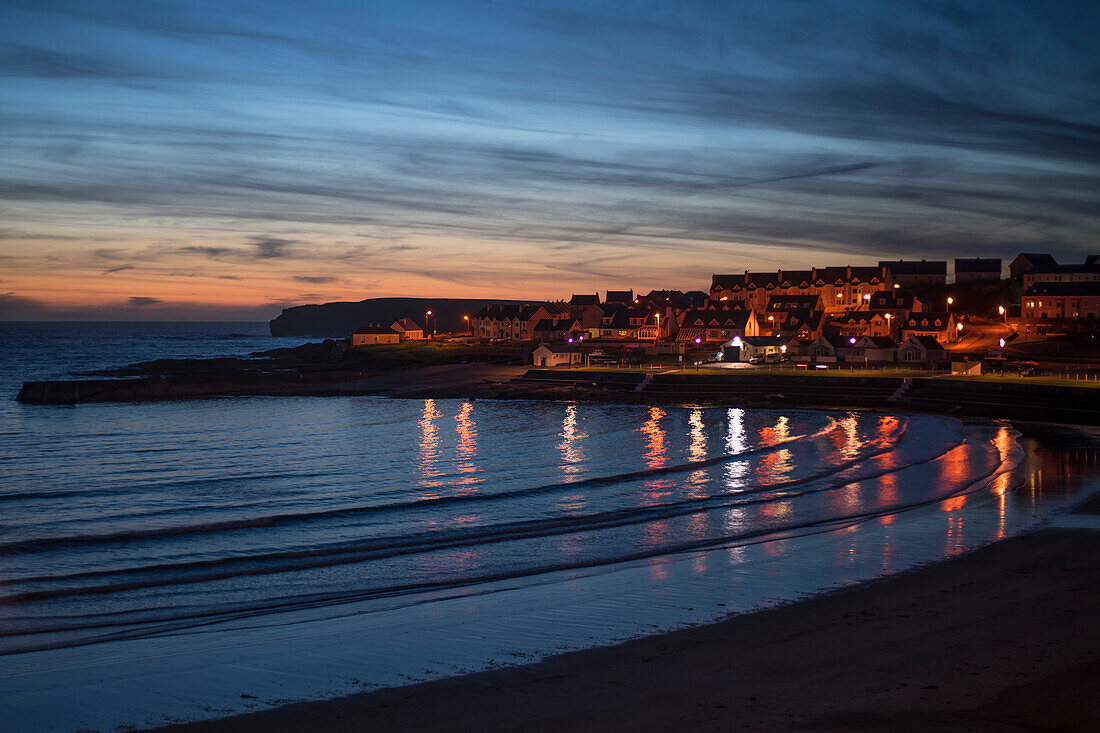 Stadtlichter, die im Wasser des Atlantiks in der Kilkee Bucht unter einem rosa Himmel während des Sonnenuntergangs reflektieren, Kilkee, County Clare, Irland, Europa