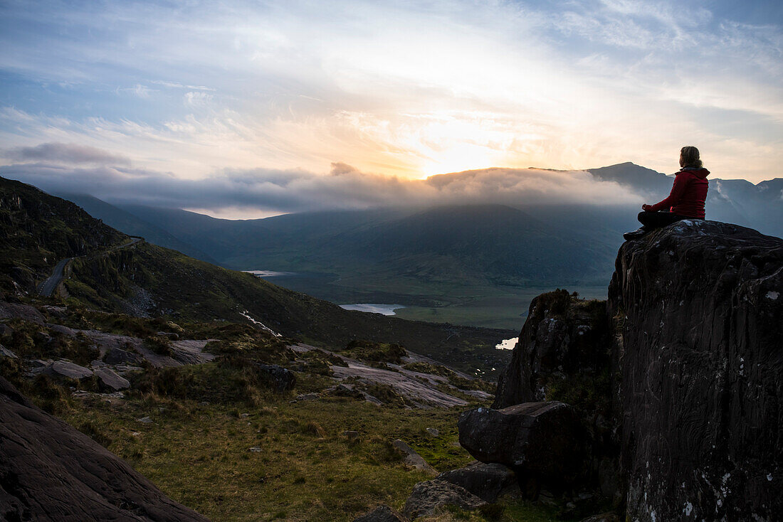 A woman sits relaxed on a rock and admires the sunset over the mountain pass with a vast panoramic landscape, seen from while walking the Dingle Way, Dingle Peninsula, County Kerry, Ireland, Europe
