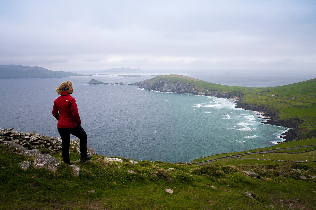 A blond woman in a red jacket and hiking boots enjoys the view of Slea Head and the Sleeping Giant of the Great Blaskets on a windy spring day seen from while walking the Dingle Way, Slea Head, Dingle Peninsula, County Kerry, Ireland, Europe