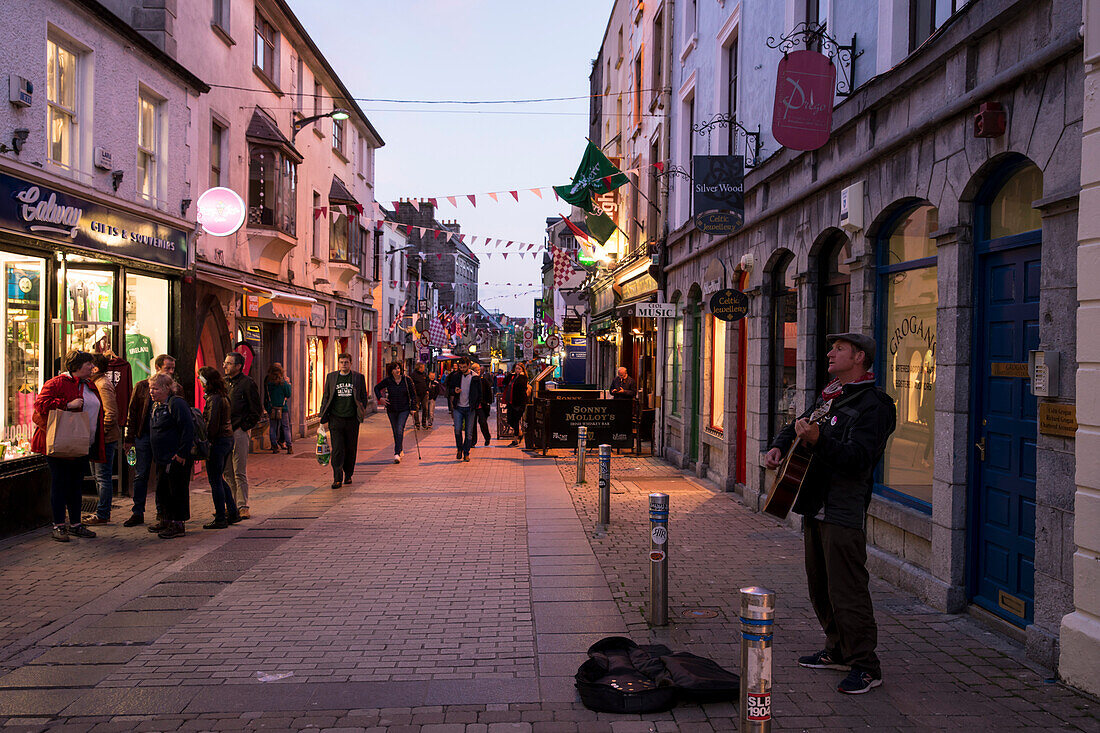Während sich die Quay Street Fußgängerzone am Abend mit Menschen füllt, singt ein Straßenmusiker aus voller Kehle und spielt seine Gitarre dazu, Galway, County Galway, Irland, Europa