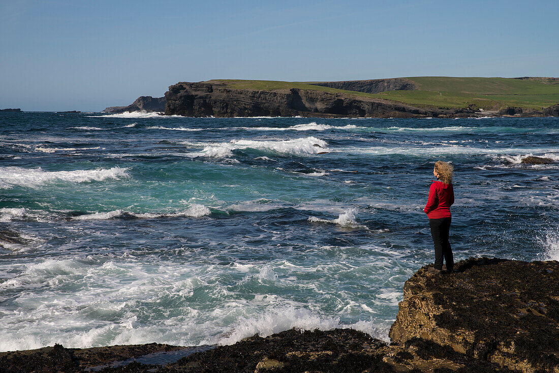 Bei einem Spaziergang an den Klippen von Kilkee betrachtet eine Frau in einer roten Jacke das aufgewühlte Meer während der Wind ihre Haare zerzaust, Kilkee, County Clare, Irland, Europa